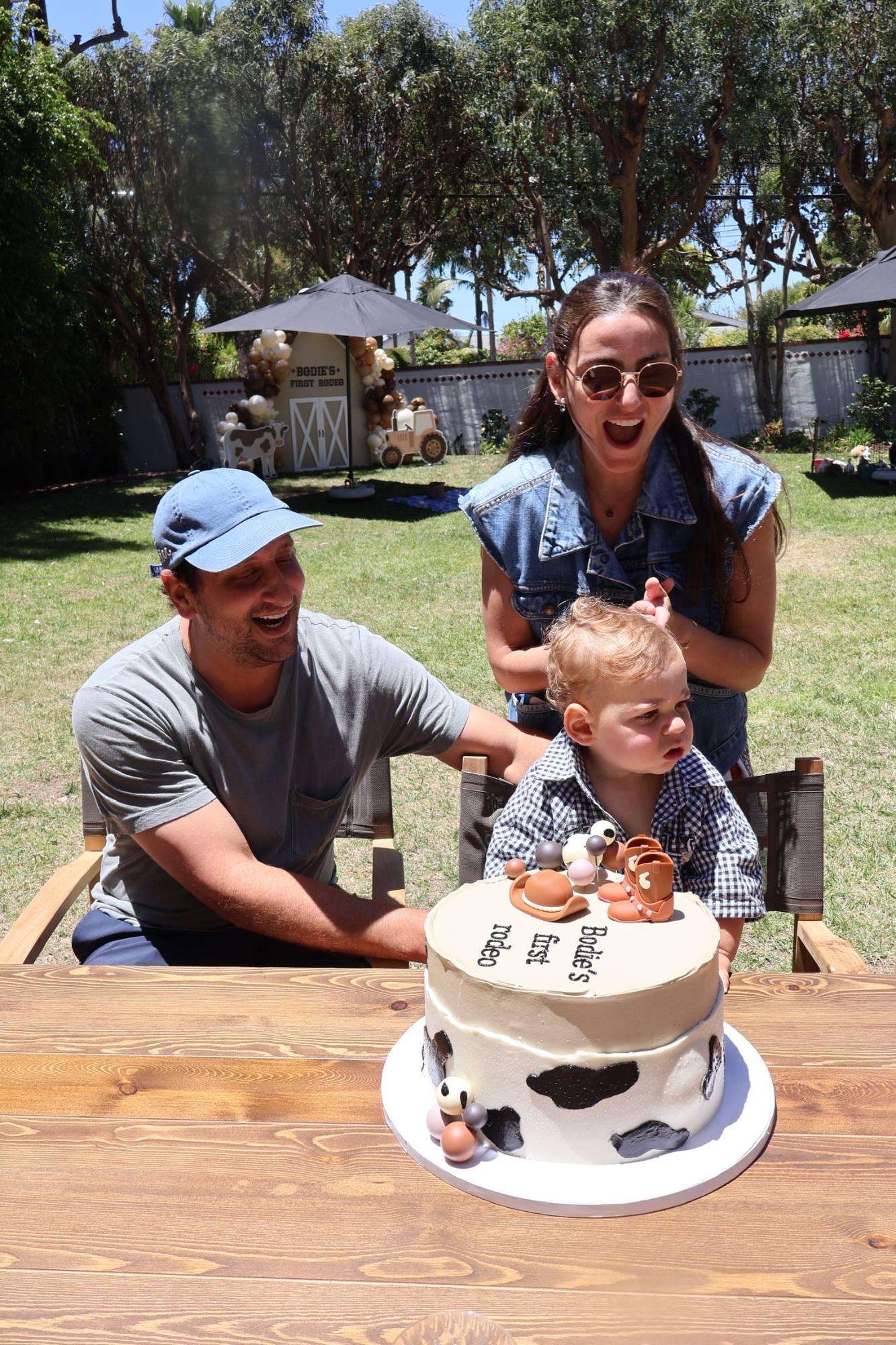 Parents laughing as their little boy sits at a garden table in front of a cow-print first birthday cake topped with tiny boots and a cowboy hat